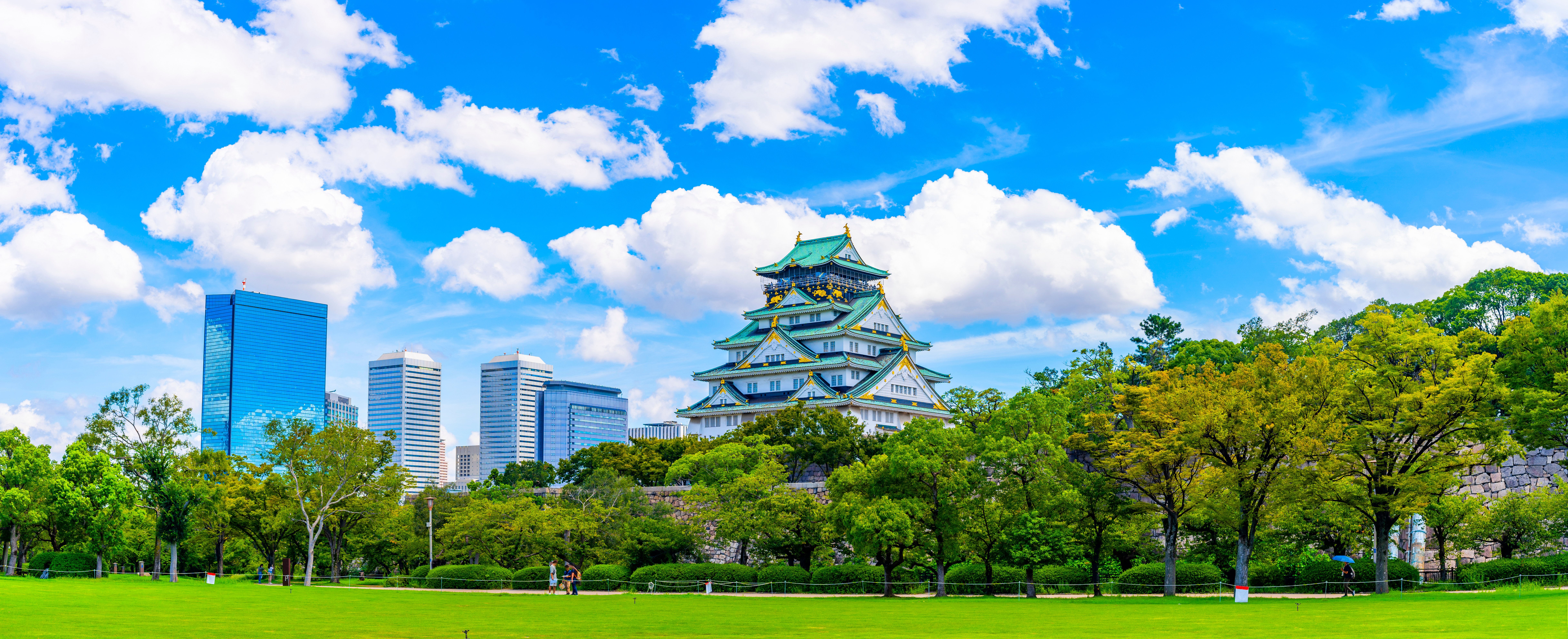Wide panoramic view of Osaka Castle from Nishinomaru Garden, surrounded by a lush green lawn and trees, with modern city skyscrapers under a bright, cloudy blue sky in the background.