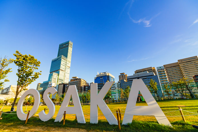 Large white 3D letters spelling "OSAKA" stand on a grassy lawn with the glass Abeno Harukas skyscraper and other city buildings under a blue sky in the background.