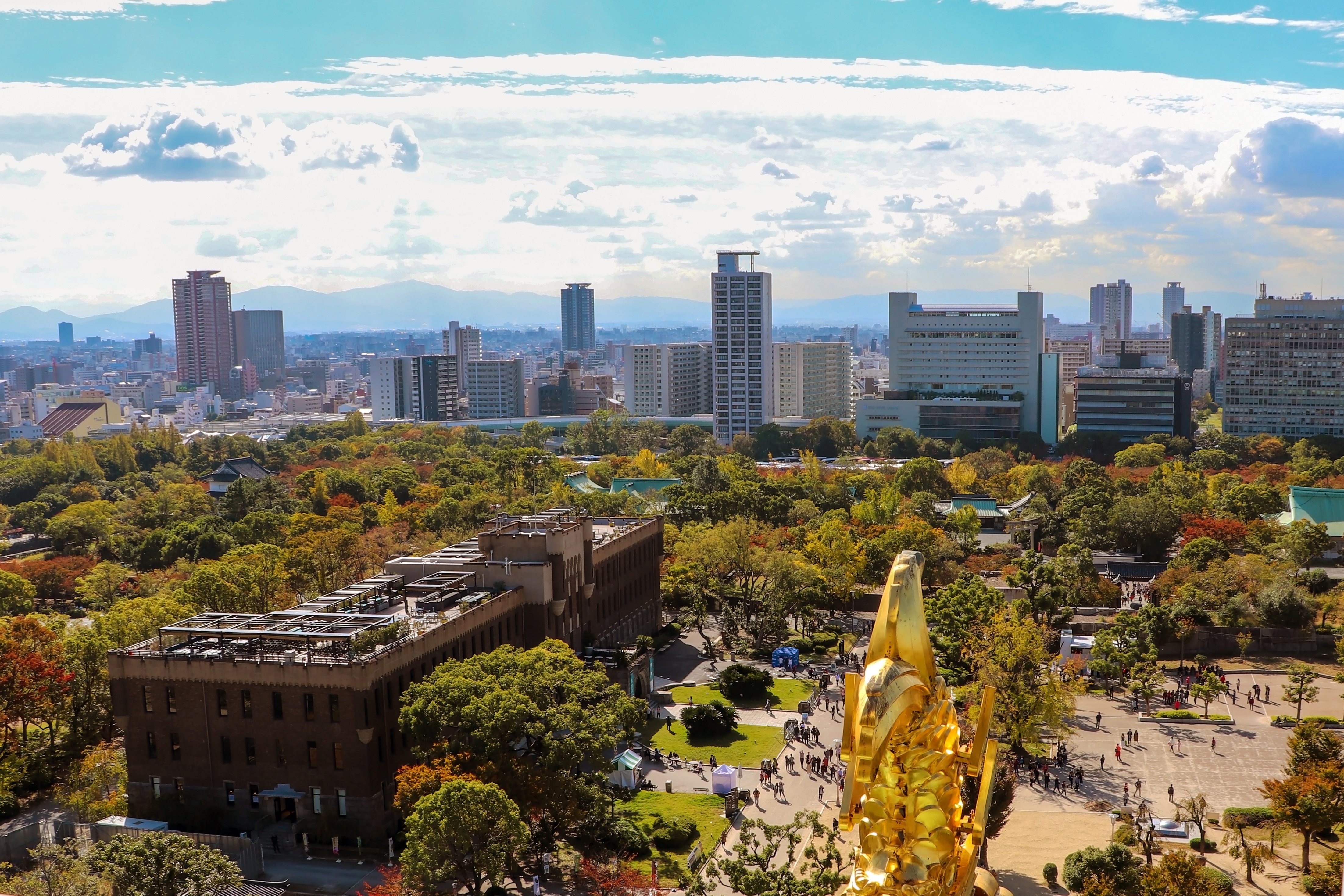 Aerial view from Osaka Castle's observation deck overlooking a golden shachihoko ornament, Osaka Castle Park, and the city skyline with Ikoma mountains in the distance