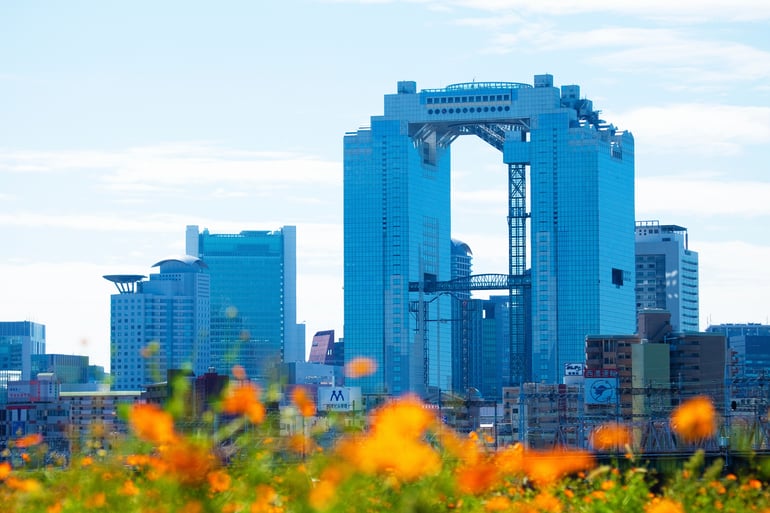 A daytime view of the modern, two-tower Umeda Sky Building in Osaka, Japan, seen rising from behind a foreground of blurred orange flowers.