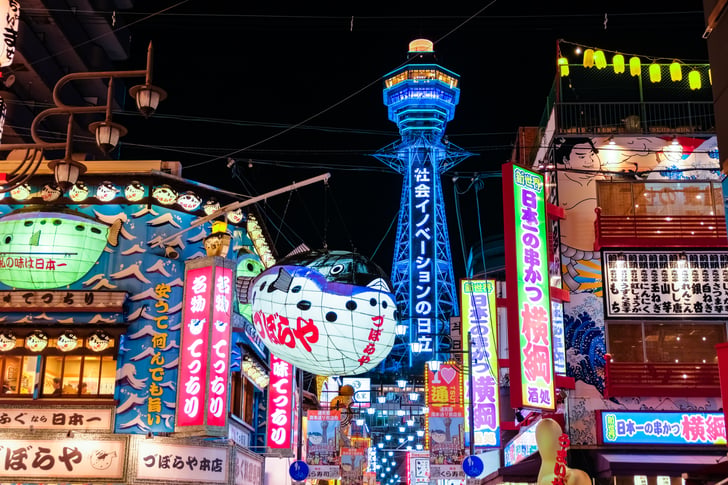 Vibrant night view of the Shinsekai district in Osaka, featuring colorful neon signs and a giant pufferfish lantern framing the illuminated blue Tsutenkaku Tower in the background.