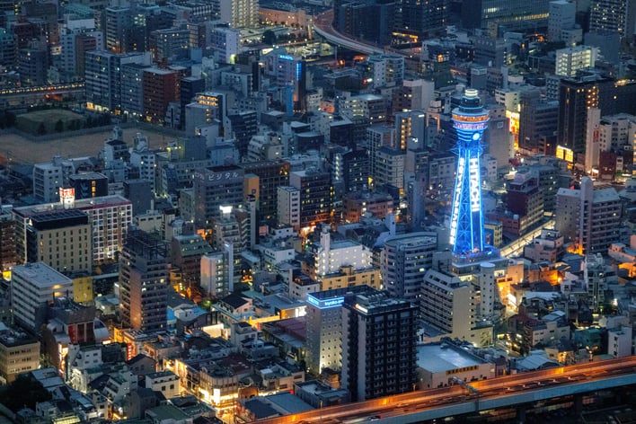 High-angle night view of the illuminated Tsutenkaku Tower glowing bright blue amidst the densely packed buildings of Osaka, Japan.
