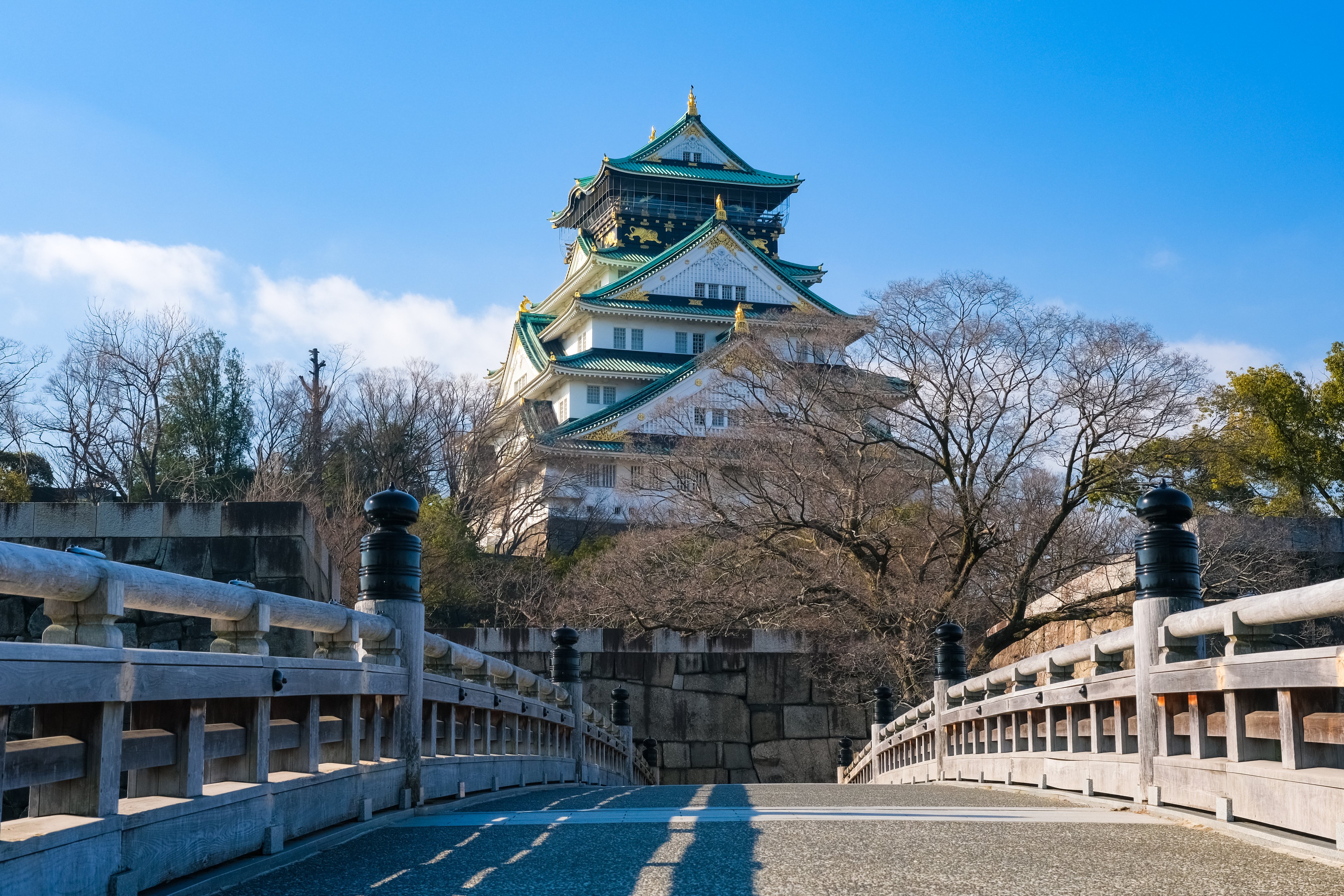 Osaka castle view from Gokurakubashi bridge, featuring the historic wooden bridge in the foreground looking up at the multi-tiered main tower surrounded by stone walls and trees under a clear blue sky..