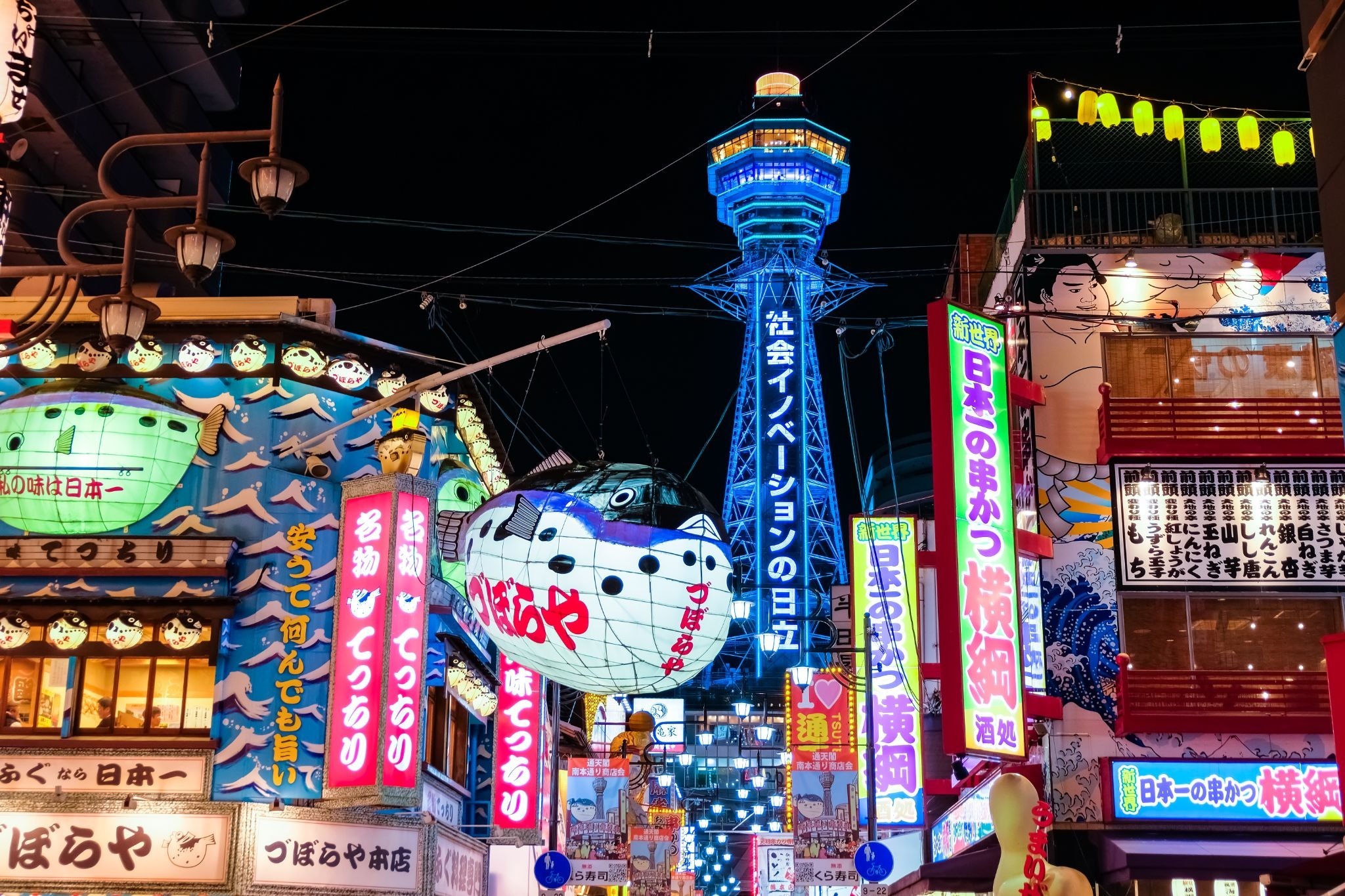 Tsutenkaku Tower illuminated at night above the colorful restaurants and neon signs of Shinsekai in Osaka.
