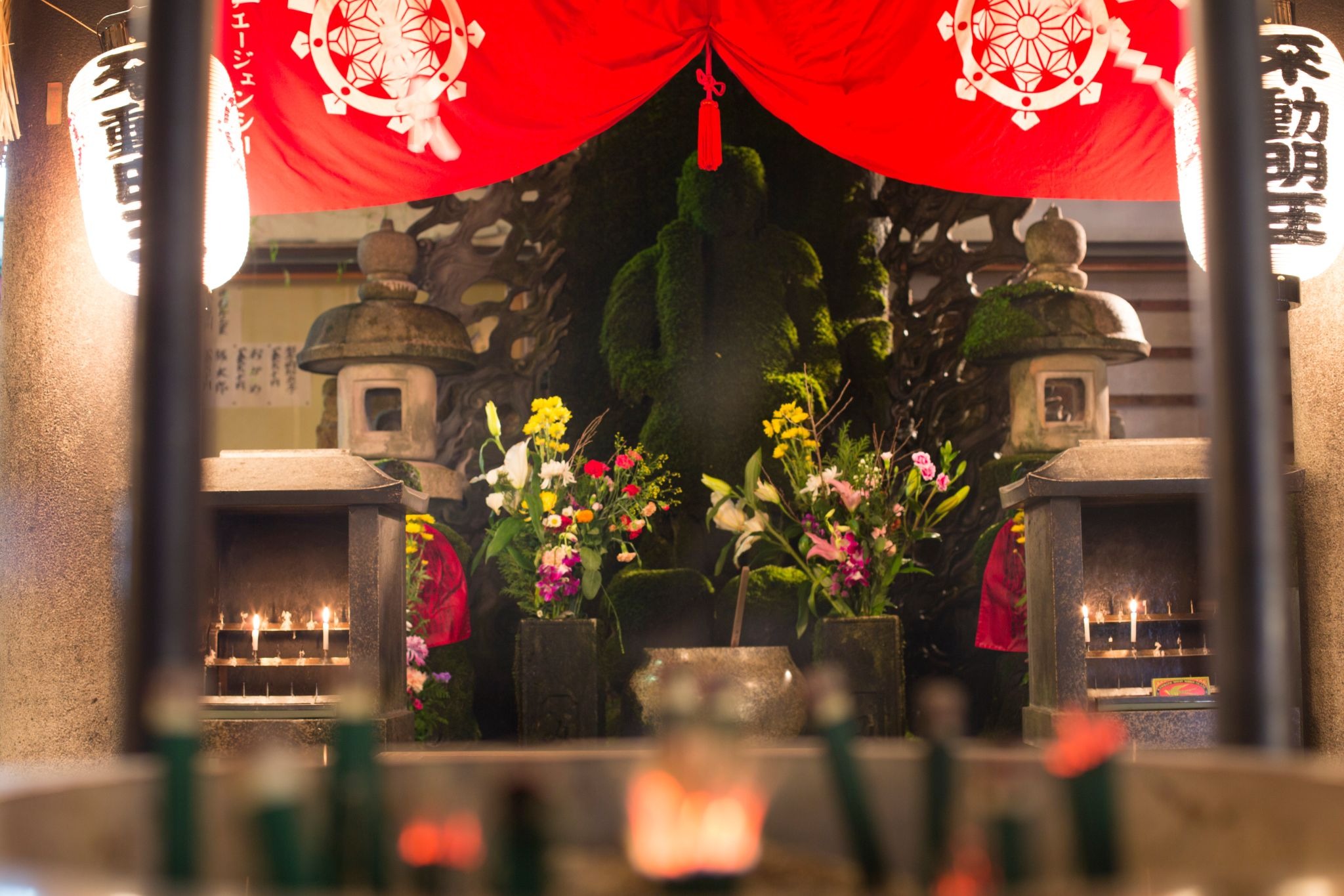 Moss-covered Fudo Myo-o statue at Hozenji Temple in Namba, Osaka, surrounded by lanterns, flowers, and offerings.